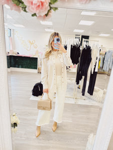 Cream wide leg frayed hem pant being worn by a woman in a boutique store location with a neutral background. Paired easily with a taupe button down, layered on top of a high neck sleeveless top and taupe fall heeled booties. Accessorized with a classic quilted crossbody bag.