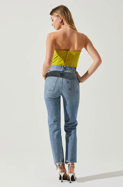 A woman in front of a light grey background showing the back of a lime green corset top. Paired with high waisted medium washed jeans with raw hem. She is wearing silver high heels. Hook & Eye back to the lime green corset.