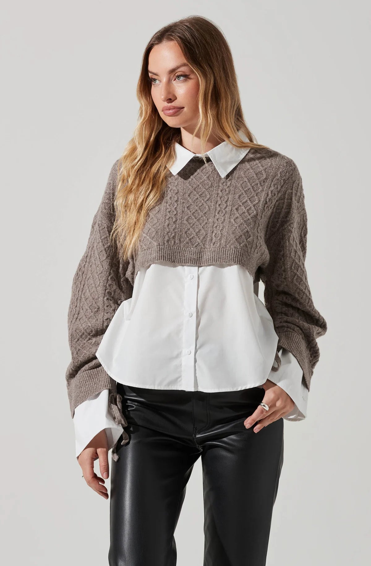 A woman wearing a mixed media top. It is a brown taupe sweater with a white button down underneath that comes as one piece. Classic collared shirt. She paired it with a faux leather high waisted pant.