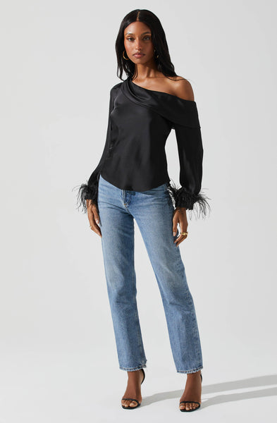 A woman in front of a light grey background wearing a black satin off the shoulder long sleeve blouse with black feather trim sleeves. Paired with a high waisted medium washed straight leg jean and a simple gold hoop and black high heels..