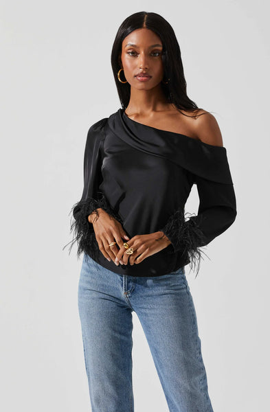 A woman in front of a light grey background wearing a black satin off the shoulder long sleeve blouse with black feather trim sleeves. Paired with a high waisted medium washed jean and a simple gold hoop.