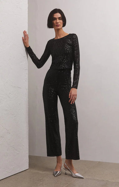 A woman in front of a white wall wearing black sequin straight leg pant. Paired with a matching black sequins long sleeve top & silver pump heels