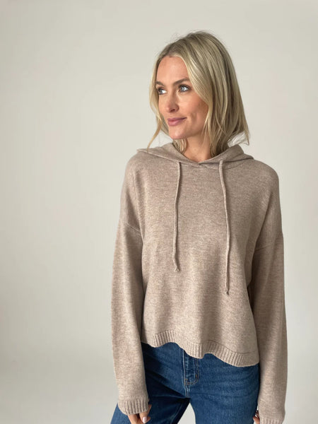 A woman in front of a white background wearing a taupe colored sweater hoodie. Paired with dark washed denim