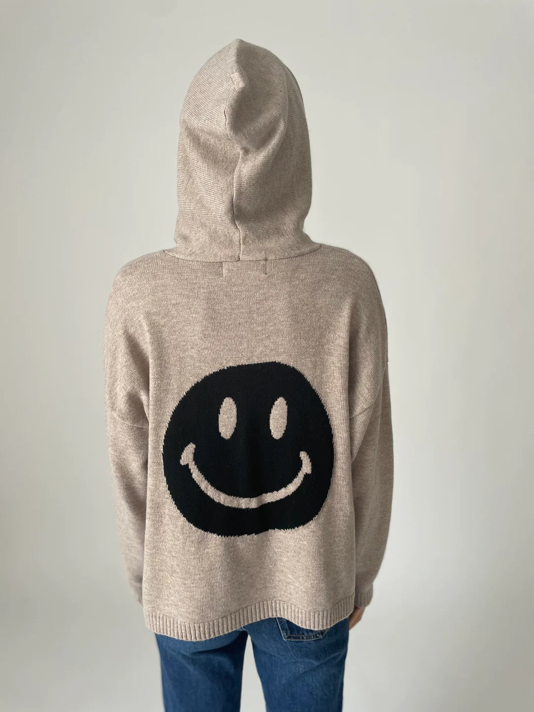 A woman in front of a white background wearing a taupe colored sweater hoodie with a black smiley face on the back of the hoodie. Paired with darker washed denim.